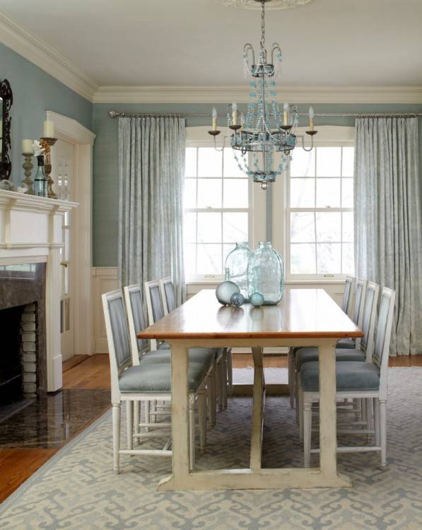 A white pleated chandelier illuminates a mahogany stained rectangular dining  table lined with seagrass dining chairs placed atop a denim blue striped rug