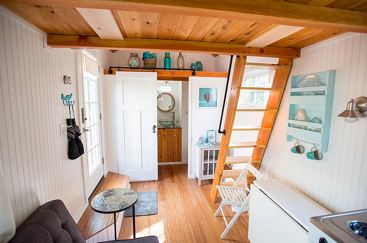 Tiny house interior with white walls, white appliances, farmhouse sink,  wood bench, potted palm, lofted bed,wood floors, oriental rug, hanging  light,